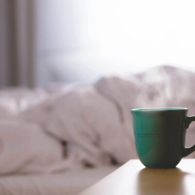 A green mug on a wooden surface with a blurred white bed in the background.