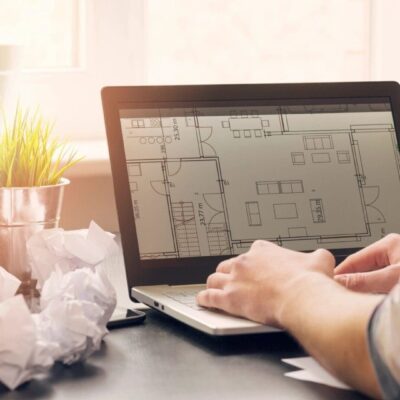 A man is working on a laptop with a blueprint in front of it.