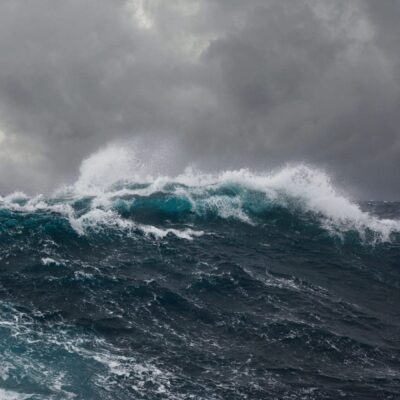 A large wave is crashing in the ocean under a cloudy sky.