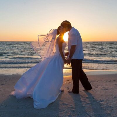 A bride and groom kissing on the beach at sunset.