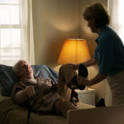 A woman helping an elderly man to his bed.