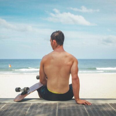 Man sitting on a boardwalk facing the ocean with a skateboard beside him.
