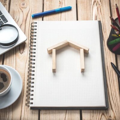 A notebook with a house model on it, a cup of coffee and a laptop on a wooden table.