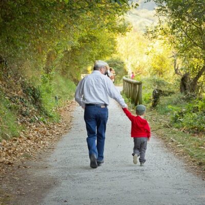 An elderly man walks hand-in-hand with a child on a tree-lined path.