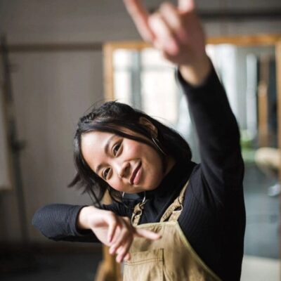 A woman in overalls is making a gesture in an art studio.