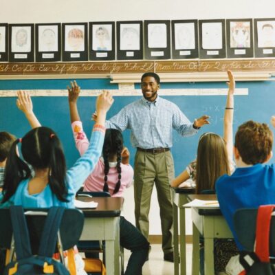 A teacher in a classroom with students raising their hands.