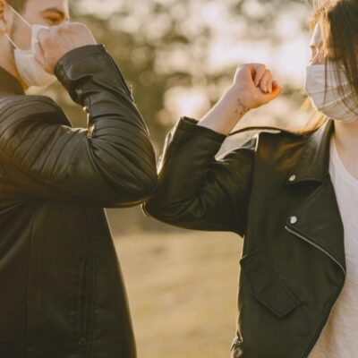 Two people greeting with elbow bumps outdoors, wearing masks.