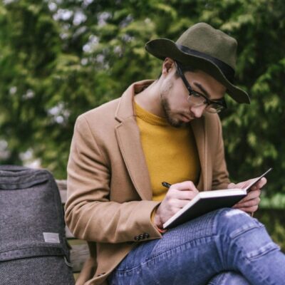 A man in a hat is sitting on a bench and writing in a notebook.