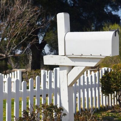 White mailbox mounted on a post beside a white picket fence.