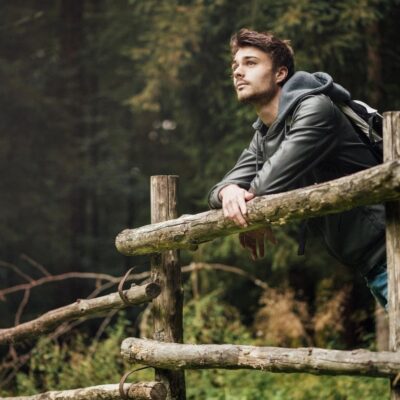 Man leaning on wooden fence in a forest, looking thoughtful.