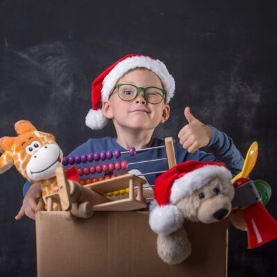 A boy in a santa hat and santa claus hat is holding toys in a cardboard box.