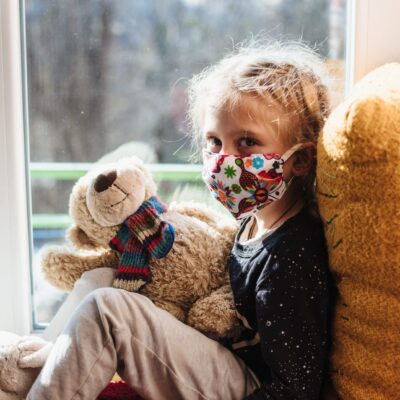 A little girl wearing a face mask sitting on a window sill with a teddy bear.
